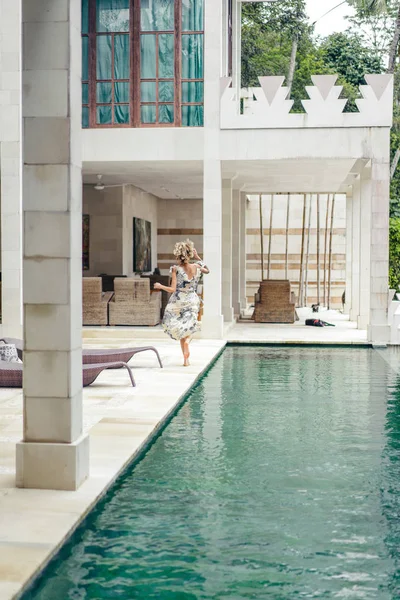 Vista trasera de la mujer en hermoso vestido corriendo cerca de la piscina en la villa - foto de stock