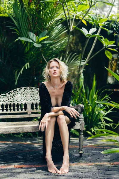 Femme blonde en vêtements noirs avec cigarette à la main reposant sur un banc sur la terrasse, ubud, bali, indonesia — Photo de stock