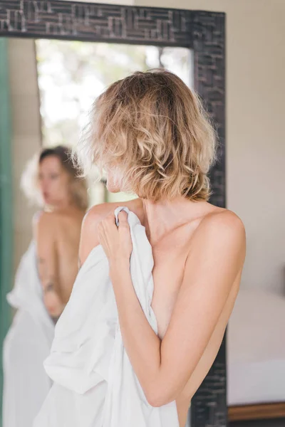 Vista parcial del espejo reflejo de la mujer rubia cubriendo el cuerpo con sábanas blancas - foto de stock