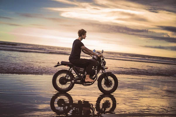 Vista lateral do homem tatuado andar de moto na praia do oceano durante o belo nascer do sol — Fotografia de Stock