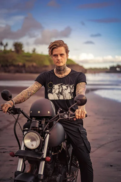 Tätowierter Biker sitzt auf Motorrad am Strand und blickt in Kamera — Stockfoto