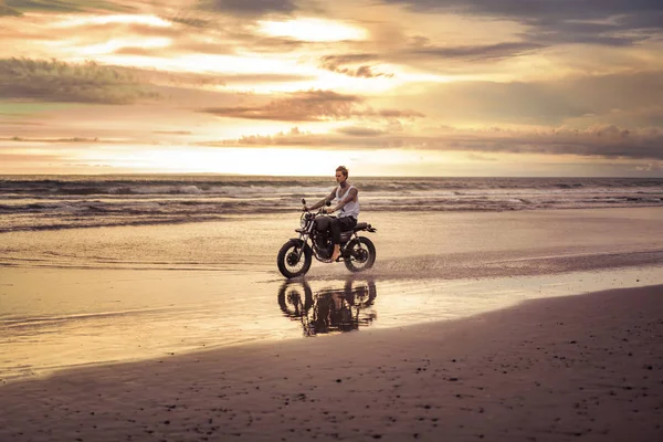 Tattooed biker riding motorcycle on ocean beach during sunset — Stock Photo