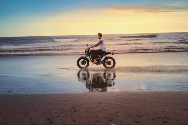 Vista lateral de motociclista tatuado andar de moto na praia do oceano durante o nascer do sol — Fotografia de Stock