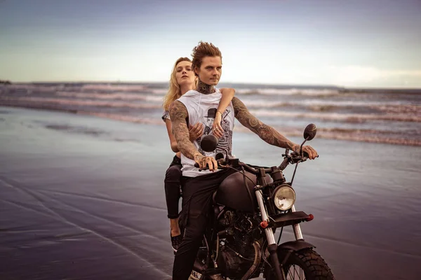 Joven elegante pareja tatuada a caballo motocicleta en la playa del océano - foto de stock