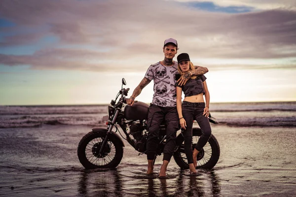 Стильний хлопець обіймає дівчину біля мотоцикла на океанічному пляжі і вони дивляться на камеру — стокове фото