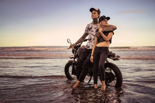 Stylish couple standing and hugging near motorcycle on ocean beach — Stock Photo