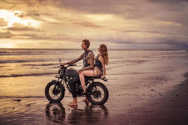 Novio y novia tatuados sentados en motocicleta en la playa durante el amanecer - foto de stock