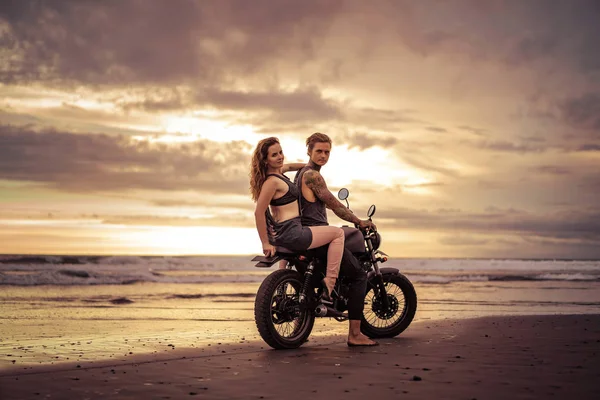 Freund und Freundin sitzen bei Sonnenaufgang auf dem Motorrad am Strand und schauen in die Kamera — Stockfoto