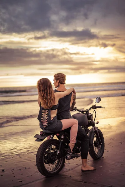 Vista trasera de novio y novia sentado en moto en la orilla del mar durante el hermoso amanecer - foto de stock