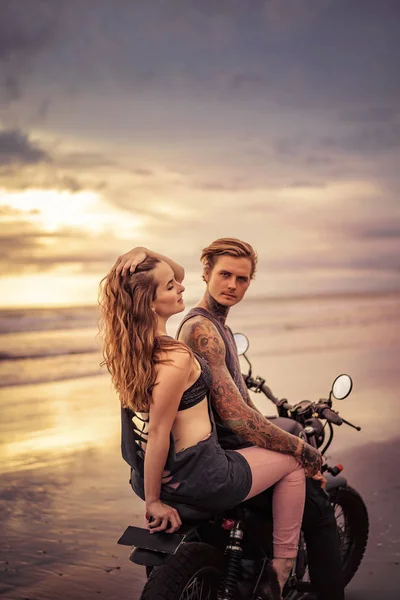 Sensual novio y novia sentado en moto en la orilla del mar durante hermosa salida del sol - foto de stock