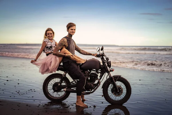 Улыбающаяся пара, обнимающаяся на мотоцикле на берегу океана во время прекрасного восхода солнца — стоковое фото