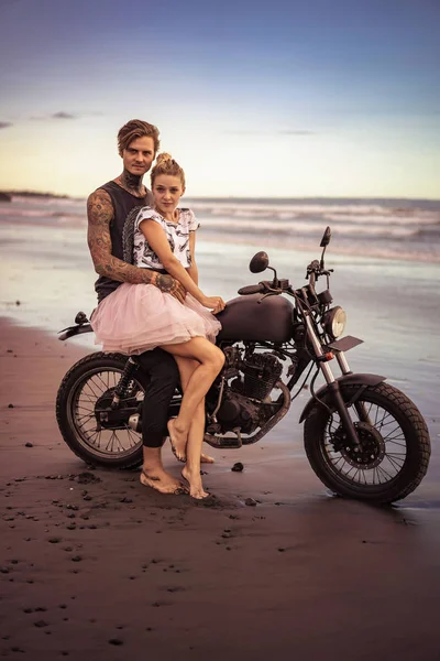 Casal abraçando na motocicleta na praia do oceano durante o belo nascer do sol e olhando para a câmera — Fotografia de Stock