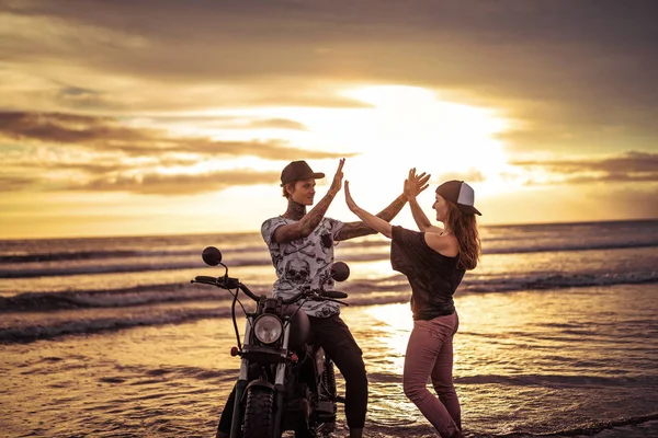 Pareja dando alta cinco cerca de la motocicleta en la playa del océano durante el amanecer - foto de stock