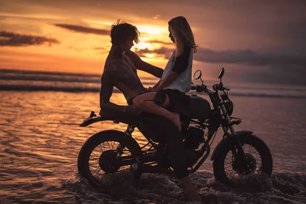Novia sentada en el novio sin camisa en moto en la playa durante la puesta del sol - foto de stock