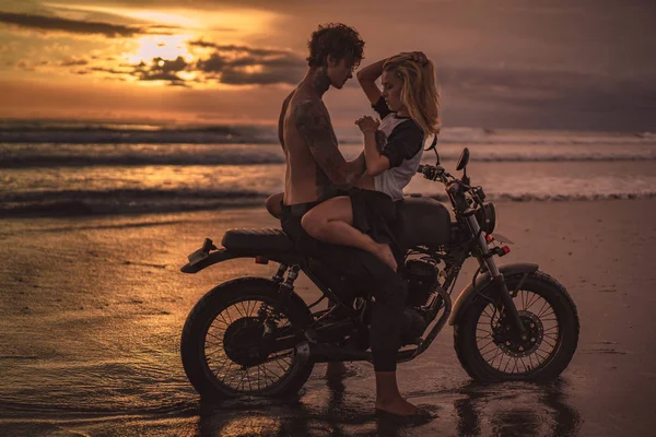 Sexy novia y novio abrazos en moto en la playa durante el atardecer - foto de stock