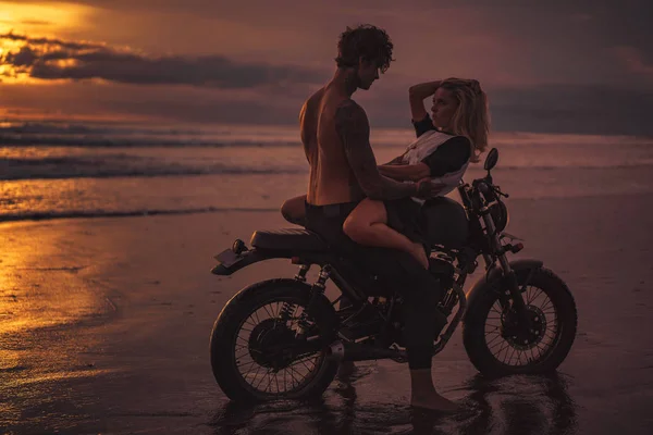 Couple séduisant câlins sur la moto à la plage pendant le coucher du soleil — Photo de stock