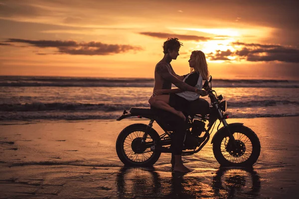 Petit ami passionné et petite amie câlins sur la moto à la plage pendant le coucher du soleil — Photo de stock