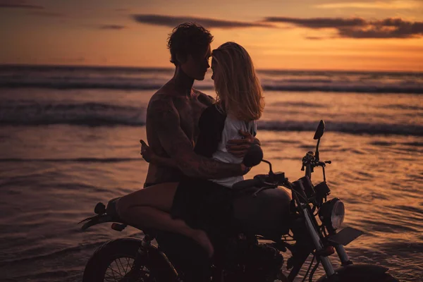 Novio apasionado y novia abrazándose en moto en la playa durante el atardecer - foto de stock