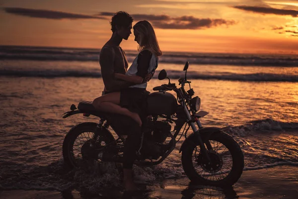 Verführerisches Paar, das sich bei Sonnenuntergang auf dem Motorrad am Strand umarmt — Stockfoto