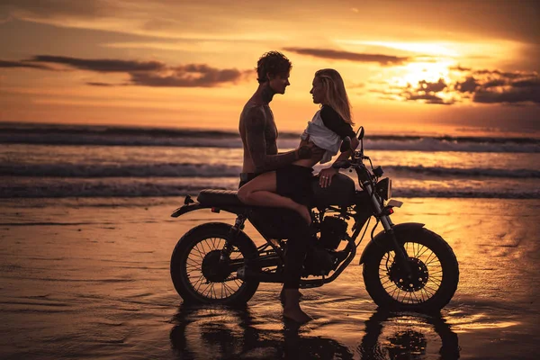Sinnliches Paar, das sich bei Sonnenuntergang auf dem Motorrad am Strand umarmt — Stockfoto