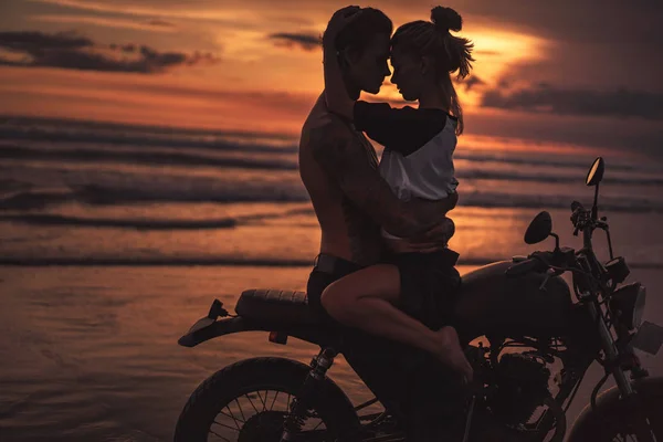 Sin camisa novio abrazando novia en moto en la playa durante el atardecer - foto de stock