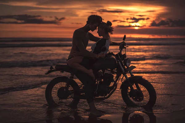 Casal apaixonado abraçando e tocando com as testas na motocicleta na praia durante o pôr do sol — Fotografia de Stock