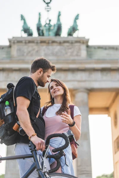 Vista de ángulo bajo de la mujer hablando con el novio con la bicicleta en frente de la Puerta de Brandenburgo en Berlín, Alemania - foto de stock