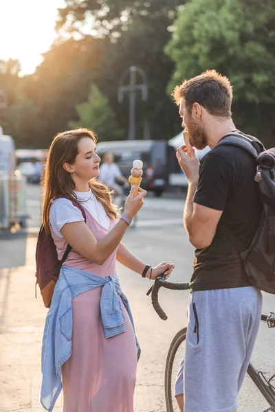 Joven con bicicleta comiendo helado mientras su novia de pie cerca de la calle - foto de stock