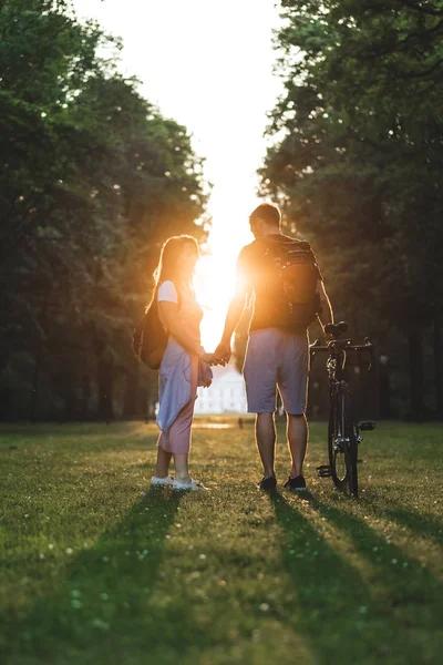 Casal com mochilas e bicicleta de pé no prado com pôr do sol para trás — Fotografia de Stock