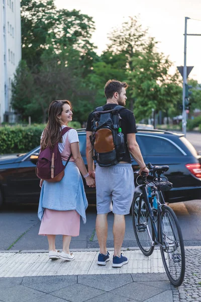 Rear view of couple with backpacks and bicycle walking at urban street — Stock Photo