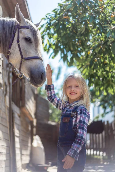 Smiling kid touching white horse at farm and looking at camera — Stock Photo
