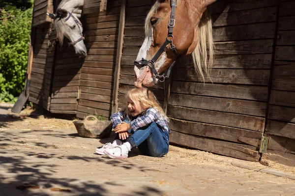 Lächelndes Kind sitzt auf dem Boden und Pferd berührt ihre Haare auf dem Bauernhof — Stockfoto