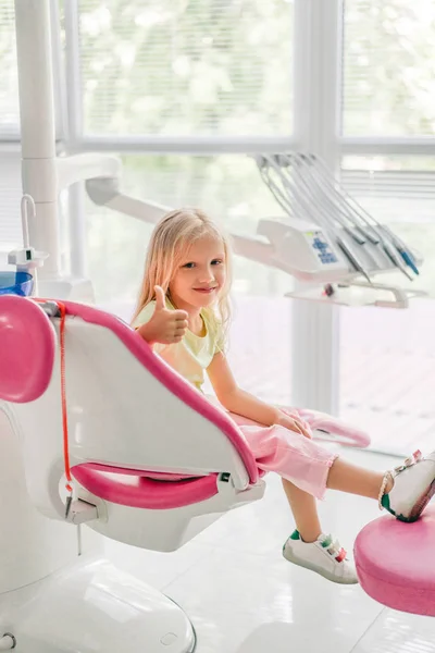 Niño sonriente mostrando el pulgar hacia arriba mientras espera al dentista en el consultorio del dentista - foto de stock