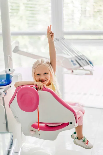 Smiling kid showing peace sign while sitting in chair at dentist office — Stock Photo