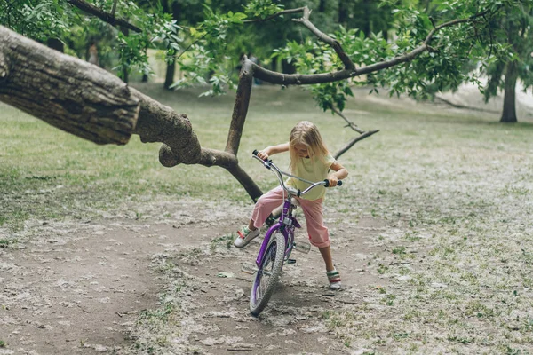 Ребенок с велосипедом стоит возле ветки деревьев в парке — стоковое фото