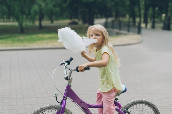 Vista lateral del adorable niño con bicicleta comiendo algodón de azúcar en el parque - foto de stock