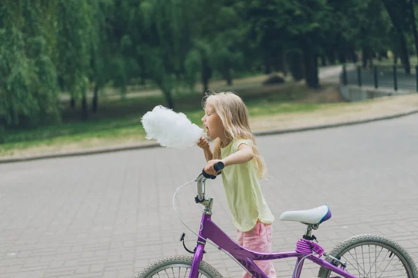 Side view of adorable kid with bicycle eating cotton candy in park — Stock Photo