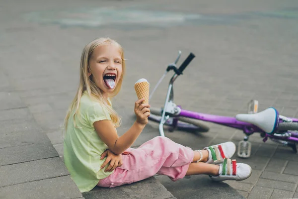 Cute child with ice cream sticking tongue out while sitting on city steps alone — Stock Photo
