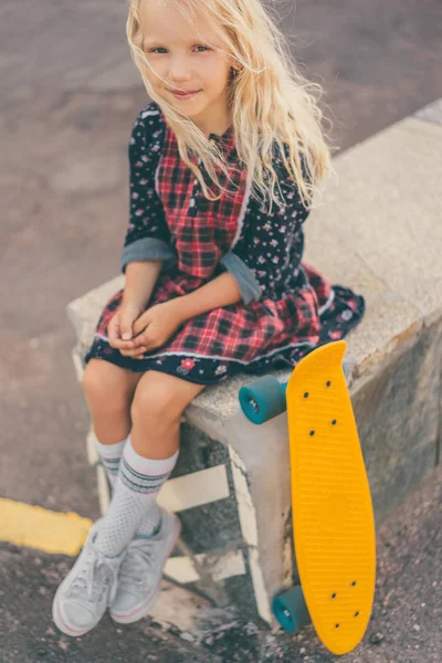 Adorabile bambino sorridente seduto vicino allo skateboard e guardando la fotocamera in strada urbana — Foto stock