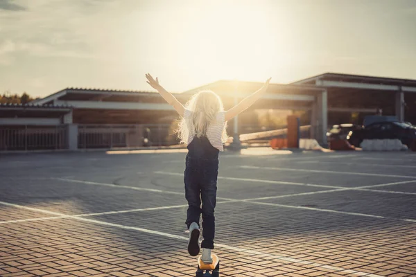 Rear view of little kid with wide arms riding on penny board at parking lot — Stock Photo