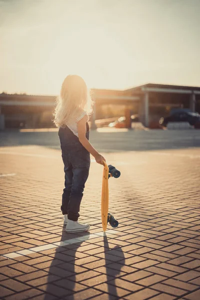 Vue arrière du petit enfant debout avec une planche de penny au parking avec coucher de soleil derrière — Photo de stock