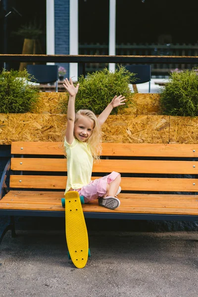Счастливый обожаемый ребенок, сидящий с широкими руками на скамейке возле скейтборда на улице — стоковое фото