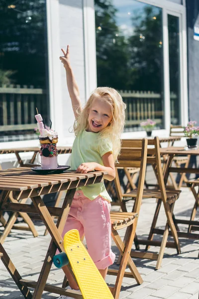 Selective focus of smiling child doing peace gesture at table with delicious dessert in cafe — Stock Photo