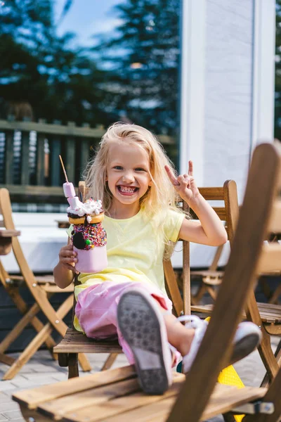 Messa a fuoco selettiva del bambino eccitato facendo gesto rock e tenendo delizioso dessert nel caffè — Foto stock