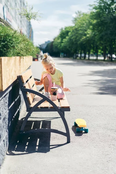 Selektiver Fokus des kleinen Kindes, das auf der Bank neben dem Skateboard auf der Straße sitzt und das Dessert betrachtet — Stockfoto