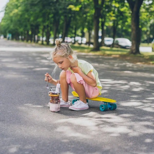 Selective focus of little child sitting on skateboard and eating dessert by spoon at urban street — Stock Photo