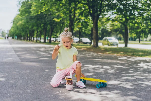 Selektiver Fokus des kleinen Kindes, das auf dem Skateboard sitzt und auf der Straße Dessert isst — Stockfoto