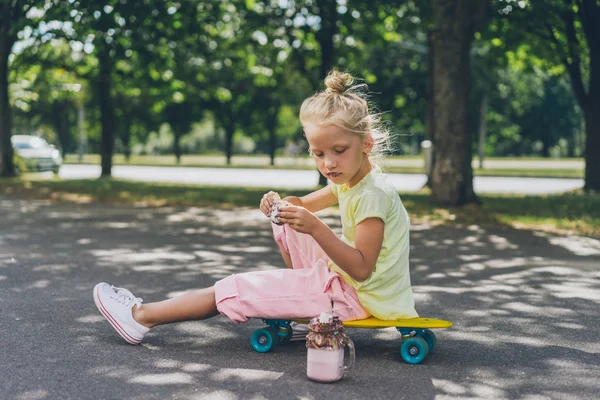 Selektiver Fokus des entzückenden Kindes, das Donut vom Dessert isst, während es auf der Straße auf dem Skateboard sitzt — Stockfoto