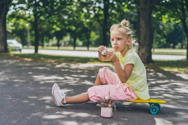 Selektiver Fokus des entzückenden Kindes, das Donut vom Dessert isst und wegschaut, während es auf der Straße auf dem Skateboard sitzt — Stockfoto