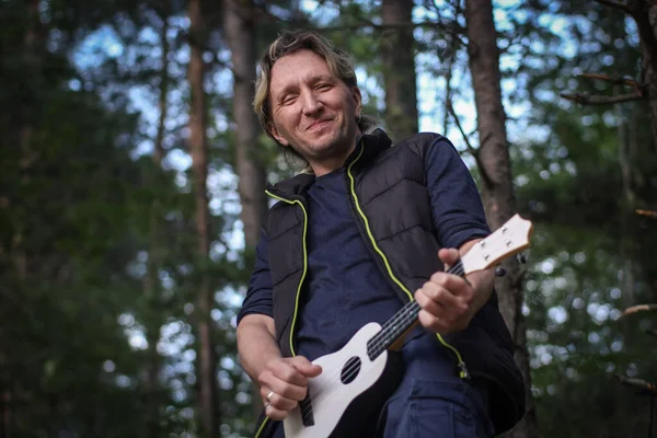 Genç adam ormanda küçük beyaz ukulele oynuyor.